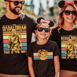 A family of three, consisting of a man, a woman, and a young girl, is wearing matching t-shirts with the same design featuring the character The Mandalorian from the Star Wars franchise.