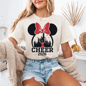 A woman wearing a white t-shirt with a graphic of a castle and a red bow, along with the text "CHEER 2016" printed on it. She is also wearing denim shorts and a beige jacket.