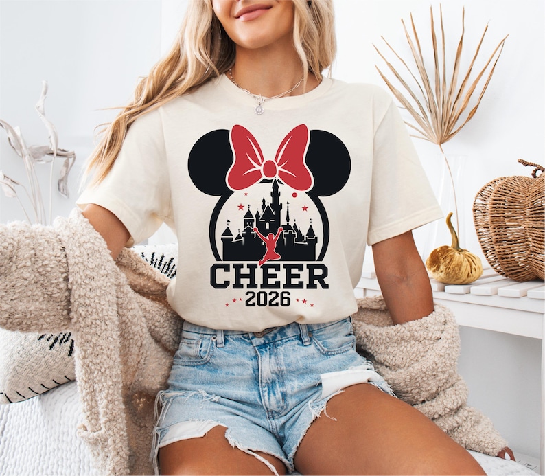 A woman wearing a white t-shirt with a graphic of a castle and a red bow, along with the text "CHEER 2016" printed on it. She is also wearing denim shorts and a beige jacket.