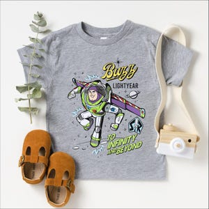 A gray t-shirt with a character from the Toy Story franchise, Buzz Lightyear, printed on it. The t-shirt also features a phrase that says "To Infinity and Beyond". The t-shirt is accompanied by a pair of brown baby shoes, a white camera strap, and some greenery.