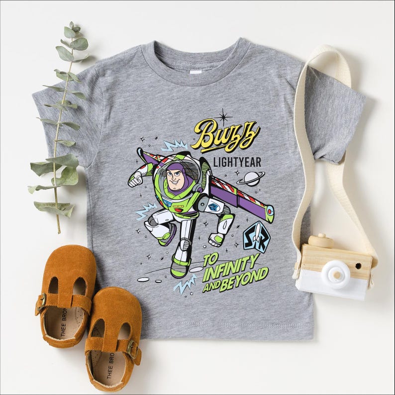 A gray t-shirt with a character from the Toy Story franchise, Buzz Lightyear, printed on it. The t-shirt also features a phrase that says "To Infinity and Beyond". The t-shirt is accompanied by a pair of brown baby shoes, a white camera strap, and some greenery.