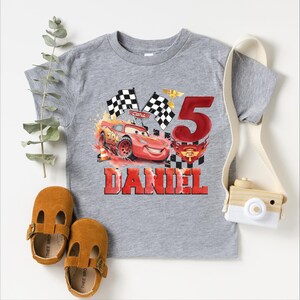A gray t-shirt with a graphic of a red car and checkered flags, along with a camera and baby shoes, suggesting a birthday or special occasion theme.