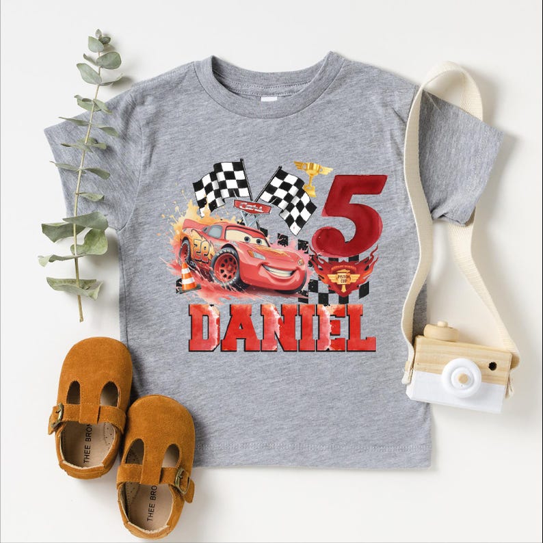 A gray t-shirt with a graphic of a red car and checkered flags, along with a camera and baby shoes, suggesting a birthday or special occasion theme.