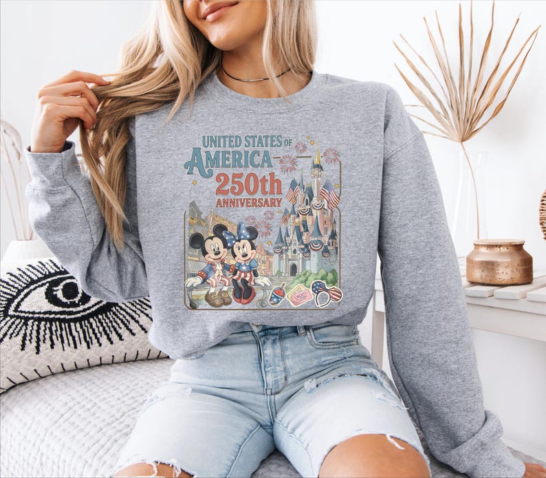 A woman wearing a gray sweatshirt with a graphic of Mickey Mouse and Minnie Mouse celebrating the United States of America 250th anniversary.