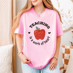 a woman wearing a pink teaching is a work of heart t - shirt