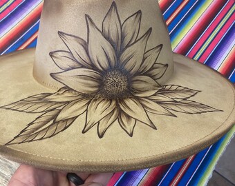 May include: A tan cowboy hat with a brown sunflower design painted on the crown.