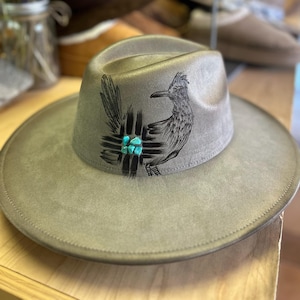 May include: A gray fedora-style hat with a wide brim. The hat features a detailed black illustration of a roadrunner bird and a turquoise stone accent. The hat is made of a soft material and has a classic, stylish design.