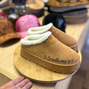 Custom Christian slippers featuring hand-burned 2 Corinthians 5:7 verse, photographed in Hat Tatt Co. workspace.
