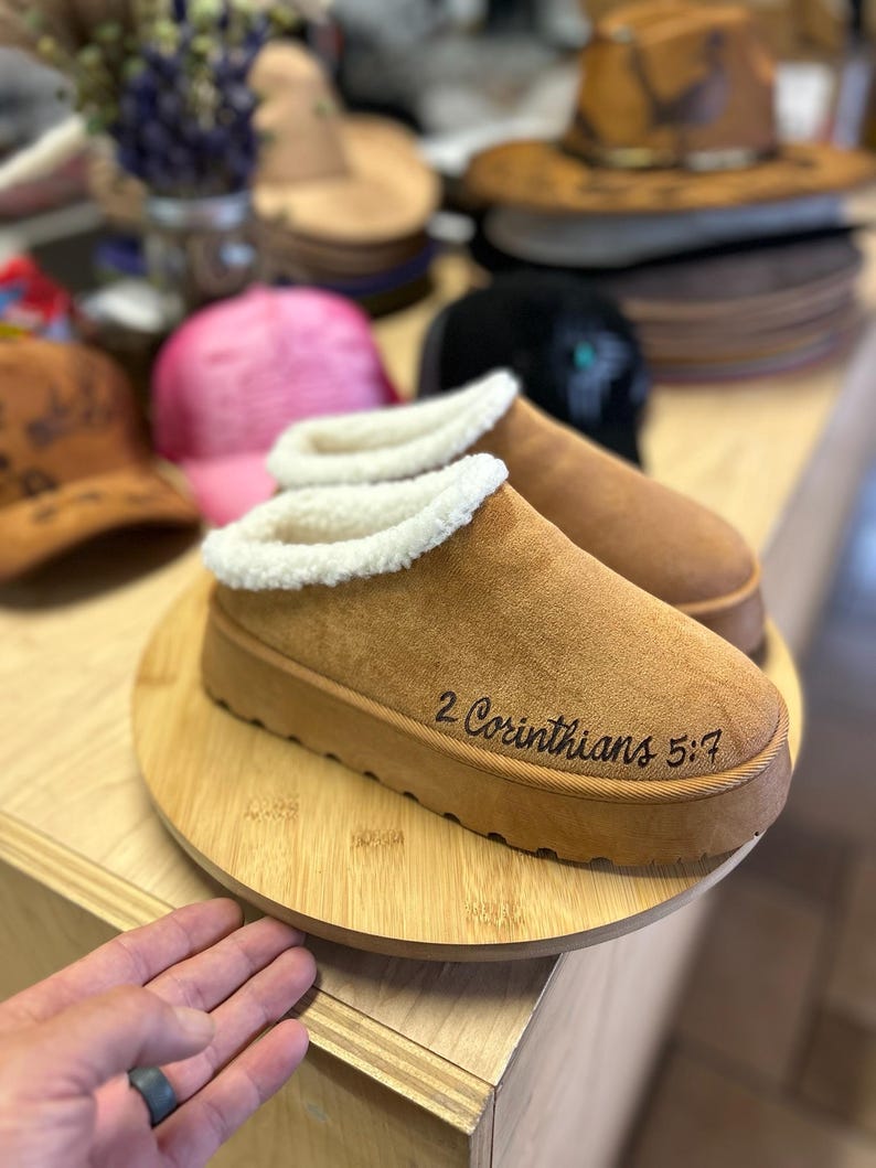 Custom Christian slippers featuring hand-burned 2 Corinthians 5:7 verse, photographed in Hat Tatt Co. workspace.