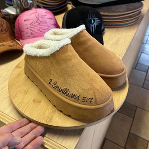 Handcrafted slippers with 2 Corinthians 5:7 engraving on side, displayed on round wood board in studio.