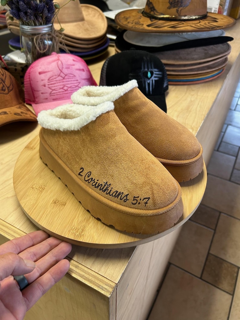 Handcrafted slippers with 2 Corinthians 5:7 engraving on side, displayed on round wood board in studio.