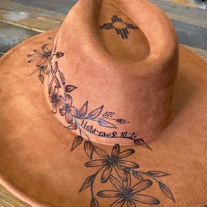 以下が含まれることがあります： 茶色のスエードハット、つば広、花柄と「ヘブライ人への手紙10:24」の文字入り。ハットのクラウンには南西部のシンボルがあります。 花柄は濃い茶色で手描きされています。