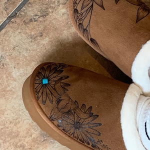 May include: A pair of brown suede slippers with a floral design and turquoise accents. The slippers have a white fleece lining.