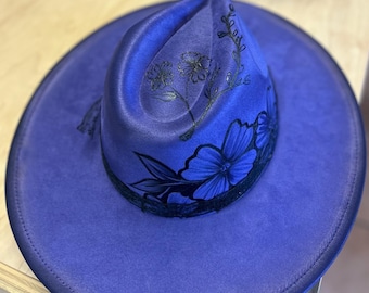 May include: A vibrant blue fedora-style hat with a wide brim. The hat features hand-painted black floral designs on the crown and brim. The hat is made of a soft material and has a dark trim.