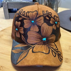 Pode incluir: Boné de beisebol castanho com um design floral. O boné apresenta flores em relevo com detalhes turquesa. A aba e as flores são delineadas em preto. O boné tem uma parte traseira de malha.