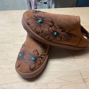 May include: A pair of brown suede slippers with a floral design and turquoise accents. The slippers have a soft, comfortable lining.