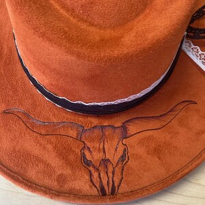 May include: A brown cowboy hat with a black and white lace trim and a black embroidered bull skull design.