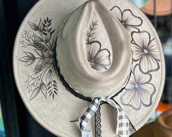 May include: A beige fedora-style hat with a wide brim, adorned with hand-painted floral and foliage designs in shades of brown and gray. The hat features a black lace band and a gingham ribbon bow.