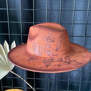 May include: A brown suede fedora hat with a black and white illustration of a roadrunner and cacti. The hat has a wide brim and a tall crown.