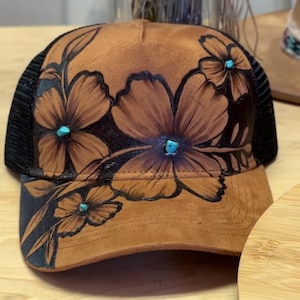Pode incluir: Boné de caminhoneiro marrom com um design floral. O chapéu apresenta um material marrom semelhante a camurça na frente com flores pintadas à mão e detalhes turquesa. A parte de trás do chapéu é de malha preta.