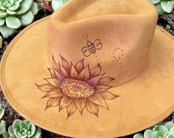 May include: A tan suede fedora hat with a sunflower and a bee design. The sunflower is brown and the bee is black and yellow.