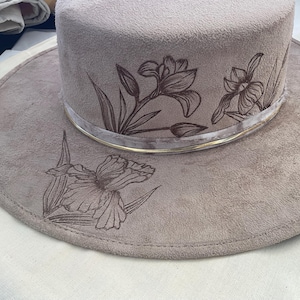May include: A light brown suede hat with a wide brim and a gold band. The hat is decorated with an embroidered floral design featuring irises.