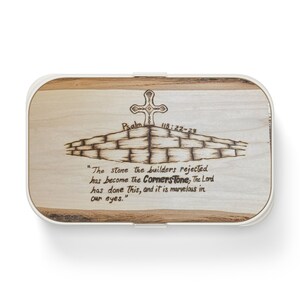 Puede incluir: Una caja de madera con una tapa blanca que presenta un diseño de madera quemada de una cruz en la parte superior de un muro de ladrillos. El texto "Salmo 118:22-23" también está quemado en la madera de la caja. El texto "La piedra que los constructores rechazaron se ha convertido en la piedra angular, el Señor ha hecho esto, y es maravilloso a nuestros ojos." también está quemado en la madera de la caja.