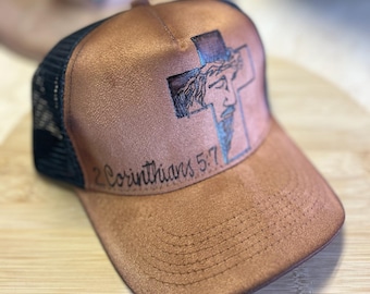 May include: Brown trucker hat with a black mesh back. The front panel features a cross design with a depiction of Jesus and the text "2 Corinthians 5:7". The hat has a curved brim and a snap closure.