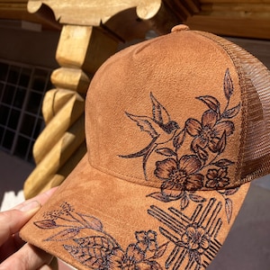 Op de afbeelding: Bruine truckerpet met een suède voorpaneel en een mesh achterkant. De pet heeft een gedetailleerd bloemen- en vogelontwerp dat in het suède is gebrand. Het ontwerp omvat bloemen, bladeren en een vogel. De pet heeft een gebogen klep.