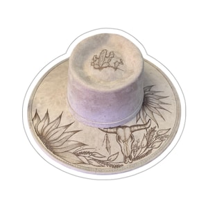 May include: A light brown cowboy hat with a white brim and a skull and floral design. The hat is decorated with a skull, cactus, and flowers.