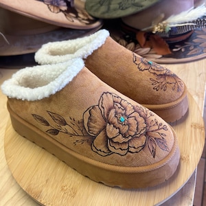 Puede incluir: Zapatillas de ante marrón con forro de piel sintética color crema. Las zapatillas presentan un gran diseño floral detallado con un acento turquesa. Otros sombreros con diseños florales están en el fondo.