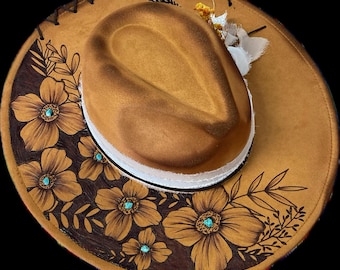 May include: A brown leather cowboy hat with a white band and a floral design. The flowers are painted in brown and have turquoise accents.