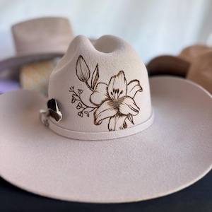 May include: A light beige cowboy hat with a floral design. The hat features a detailed, dark brown lily and leaf pattern on the crown. A small, decorative silver clasp is on the side. Other hats are visible in the background.