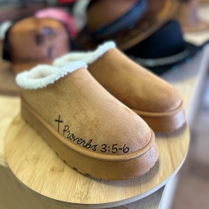 Puede incluir: Un par de zapatillas sin cordones de color beige con suela gruesa y ribete interior blanco y esponjoso. El texto "Proverbios 3:5-6" está escrito en escritura negra en el lateral. Las zapatillas se exhiben sobre una superficie de madera.