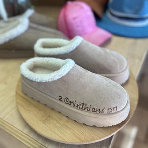 May include: Tan slip-on slippers with a white faux fur lining and a thick sole. The text "2 Corinthians 5:7" is printed on the side of the slipper. Other hats are visible in the background.