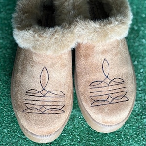 May include: A pair of tan suede slippers with a faux fur trim. Each slipper features a black embroidered design on the toe. The slippers have a thick sole and are designed for indoor wear.