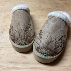 Puede incluir: Un par de zapatillas de ante marrón con forro de piel sintética. Las zapatillas tienen un diseño de girasol con detalles turquesa.