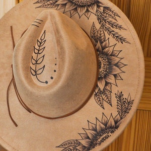Puede incluir: Un sombrero de fieltro marrón con un diseño de girasol negro. El sombrero tiene una ala ancha y una banda de cuero marrón.