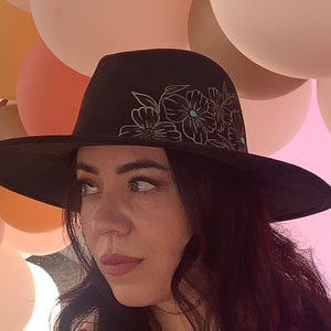 May include: Black fedora hat with a wide brim, adorned with a floral design in white and turquoise accents. The hat has a classic shape and is a stylish accessory.