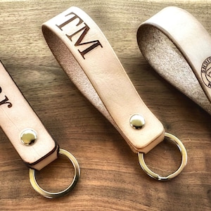 May include: Three leather keychains with metal key rings. The keychains are personalized with the names "Edmar", "TM", and a logo that says "Erbium Designs".