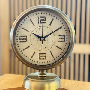 May include: A round table clock with a metallic gold frame and a circular base. The off-white clock face features black numerals at the 12, 3, 6, and 9 positions, with black tick marks for other hours and black hour, minute, and second hands.