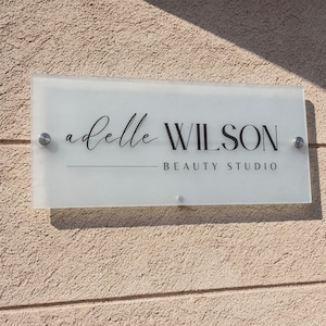 May include: A rectangular glass sign with the text "adelle WILSON BEAUTY STUDIO" in black lettering. The sign is mounted on a textured beige wall with silver hardware.
