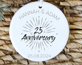 May include: A white ceramic ornament with the text "HANNAH & ADAM" and "25 Anniversary" in black. The ornament features a starburst design and the date "25.08.2024". A white ribbon is attached for hanging.