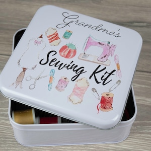 Può includere: Scatola da cucito in metallo bianco con un design colorato che presenta una macchina da cucire, bobine di filo, forbici e spilli da balia. Il coperchio ha la scritta "Grandma's Sewing Kit" stampata.