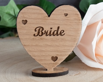 May include: Wooden heart-shaped table card with the word "Bride" engraved on it. The card is on a wooden base and has three small hearts engraved around the edges.