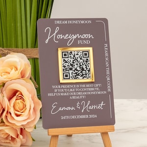 May include: A grey card with a gold frame around a QR code. The text on the card reads "Dream Honeymoon Honeymoon Fund" and "Please scan the QR code". The text also reads "Your presence is the best gift! If you'd like to contribute, help us make our dream honeymoon a reality! Eamonn & Harriet 24th December 2024".