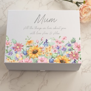 Peut inclure: Une boîte cadeau blanche avec un motif floral et le mot "Mum" en écriture manuscrite. La boîte présente une bordure de style aquarelle de tournesols, de fleurs roses, violettes et bleues. Le texte en dessous indique "All the things we love about you with love Ava & Alex."