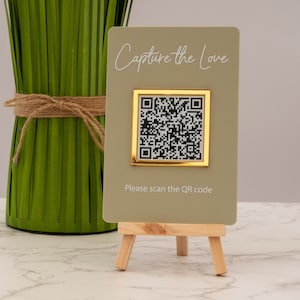 May include: A light green card with the text "Capture the Love" and a gold framed QR code. The card is on a small wooden easel.