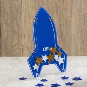 Op de afbeelding: Een blauwe raket van acryl met een transparante standaard. De raket heeft een wit naamplaatje met de tekst "Ollie" en is versierd met gouden en witte glittersterren.