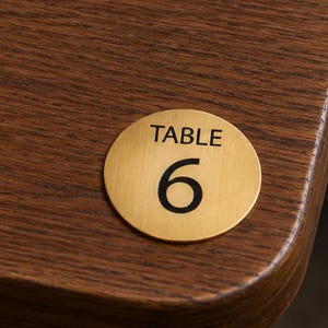 May include: A circular gold-toned metal table number marker with the text "TABLE 6" in black, positioned on a dark brown wood-grain surface next to a folded beige napkin, a silver fork, a silver knife, and a clear glass tumbler.